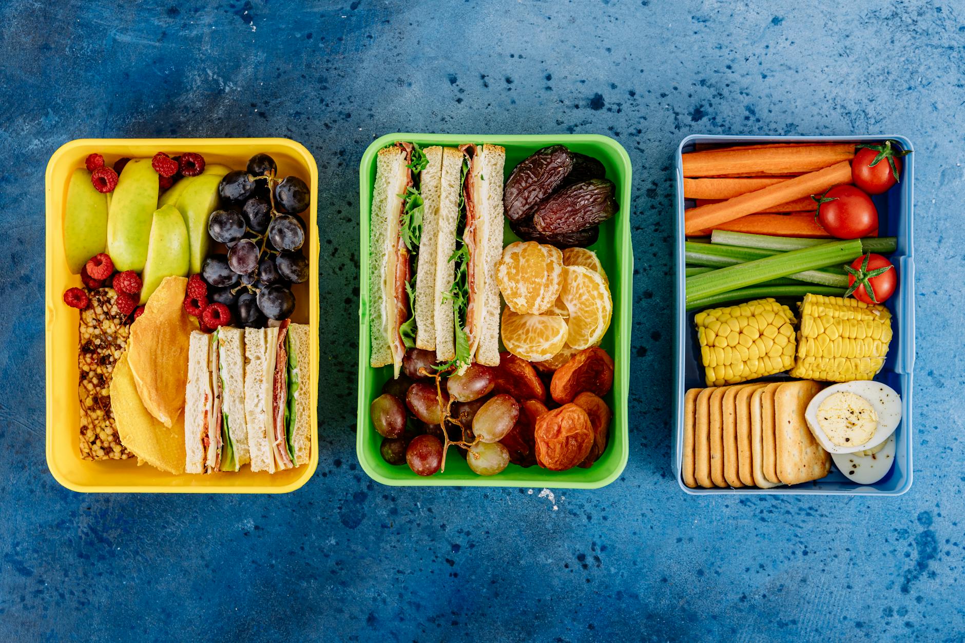 Kolorowe lunchboxy z kanapkami, owocami i warzywami na niebieskim tle